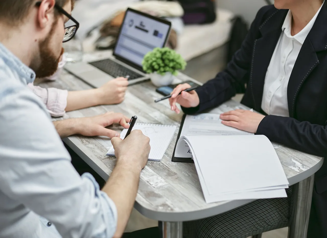 Zwei Personen sitzen an einem Tisch mit Unterlagen und Laptop bei einem Beratungsgespräch zur Beantragung eines Bildungsgutscheins