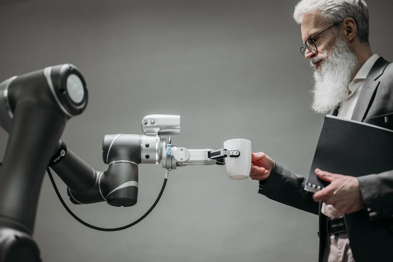 Älterer Geschäftsmann im Anzug nimmt eine Tasse von einem Industrierobotikarm entgegen – Symbol für KI-Automatisierung und die Bedrohung von Arbeitsplätzen durch künstliche Intelligenz
